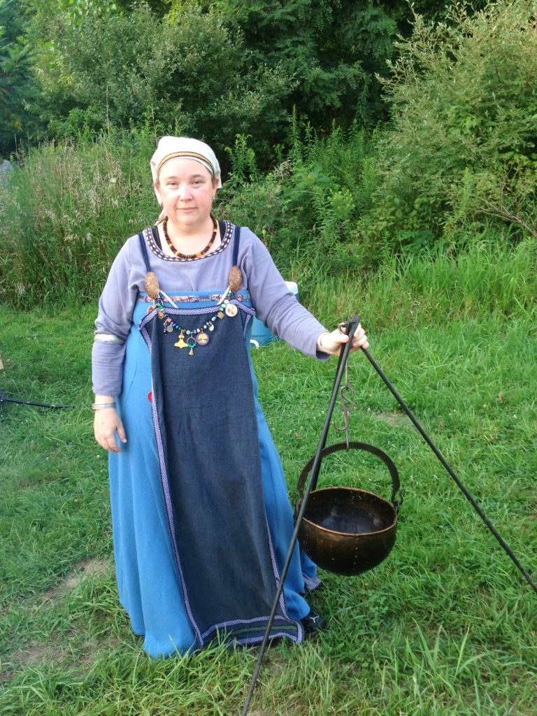 Reyni-Hrenfa (Jen), wearing her blue viking age garb, stands next to an iron tripod that has an empty viking age kettle on it.