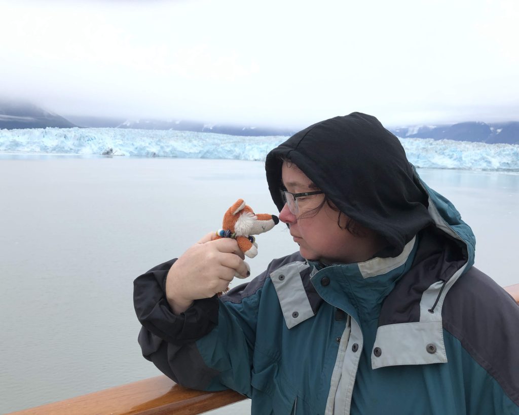 Olaf the stuffed fox, and Jen touch noses while leaning against the railing of a cruise ship, in front of an iceberg.